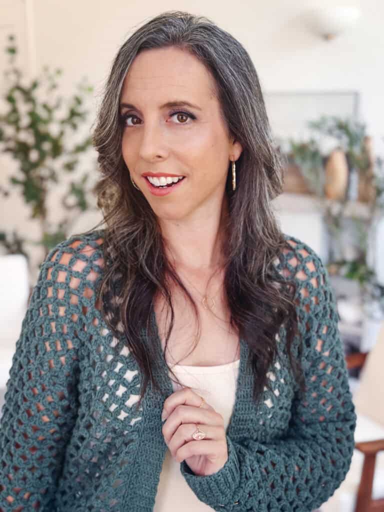 Woman with long wavy brown and gray hair wearing a green Circle Leaf Lace Crochet Cardigan pattern and cream top, standing indoors with plants and decor visible in the background.