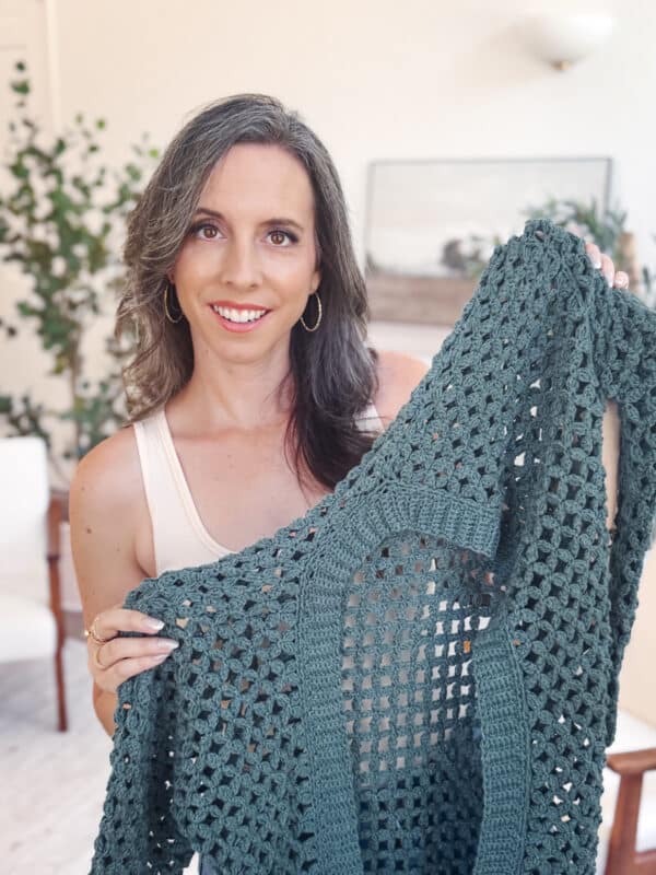A woman with long brown hair holds up a green crocheted garment, showcasing a beautiful crochet cardigan pattern in a bright indoor setting with plants and furniture in the background.