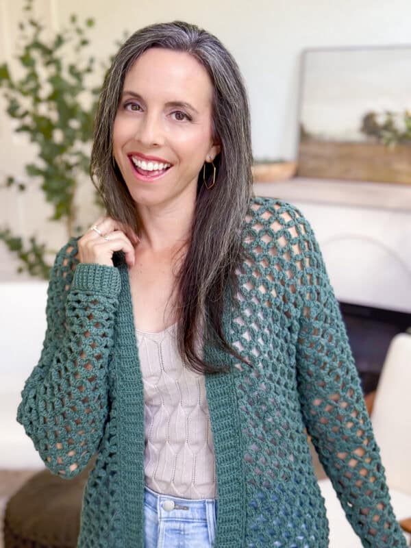 Woman with long gray-streaked hair smiling, wearing a green crochet cardigan pattern over a beige top and jeans, standing indoors in a cozy living room setting.