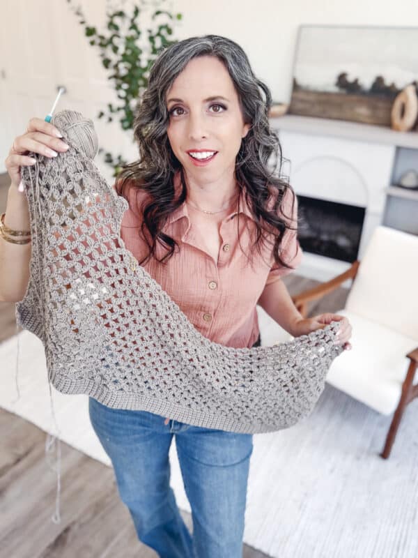 A woman with wavy gray hair holds up a partially crocheted gray garment, working on a Circle Leaf Lace Crochet Cardigan pattern, standing in a living room with a chair, rug, and plant in the background.