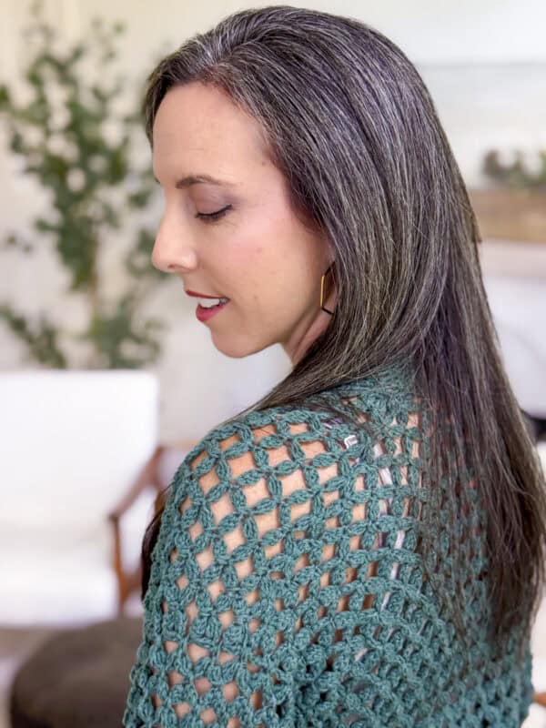 A woman with long, straight, brown and gray hair is shown in profile, wearing a green crochet cardigan pattern top indoors with blurred furniture and plants in the background.