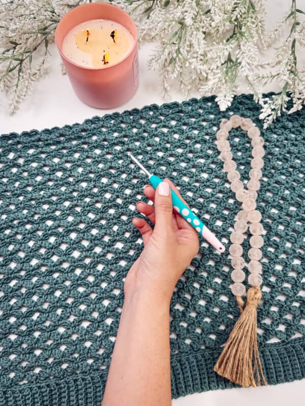 A hand holds a crochet hook over a green crocheted mat, with a strand of beads, a pink candle, and snowy foliage nearby&mdash;perfect inspiration for your next crochet cardigan pattern.