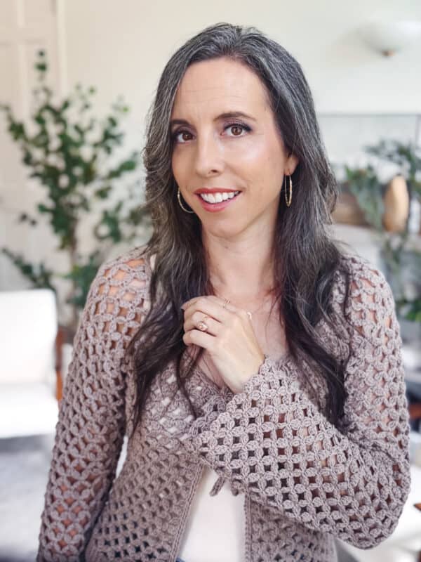 Woman with long dark hair and hoop earrings wearing a beige Circle Leaf V-Neck Crochet Cardigan pattern, standing indoors with greenery in the background, facing the camera and smiling.