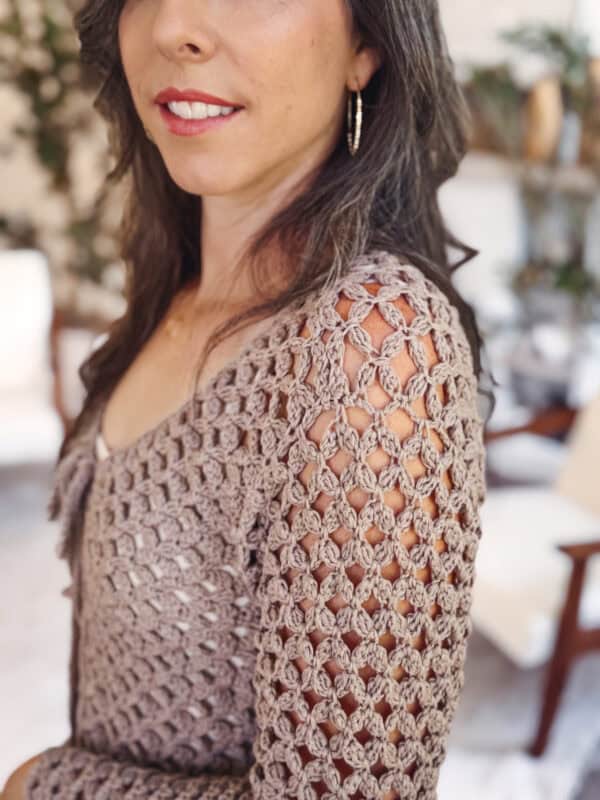 A woman wearing a taupe crochet sweater inspired by the Circle Leaf Lace Crochet Cardigan pattern, with hoop earrings, indoors amid blurred furniture and plants in the background.