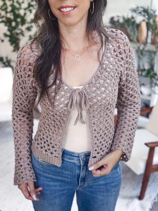A woman wears a light brown Circle Leaf Lace Crochet Cardigan over a beige top and blue jeans, standing indoors with plants and furniture in the background.