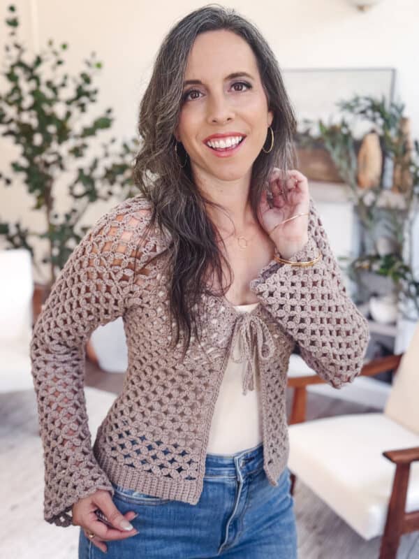 Woman with long brown hair wearing a Circle Leaf Lace Crochet Cardigan pattern over a light top and blue jeans, standing indoors with plants and chairs in the background.