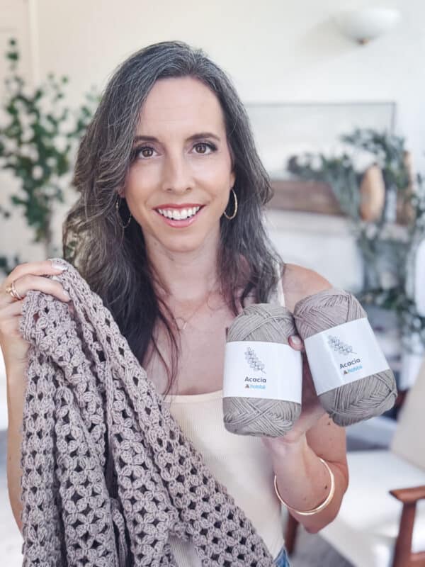 A woman holds a crocheted blanket in one hand and two skeins of Acacia yarn in the other, smiling at the camera in a well-lit room, wearing her finished Circle Leaf Lace Crochet Cardigan pattern.