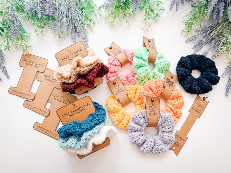 Crocheted scrunchies in various colors are arranged with brown cardboard tags on a white surface, surrounded by sprigs of purple and green artificial flowers—perfect accessories to pair with your favorite crochet nesting trays pattern.