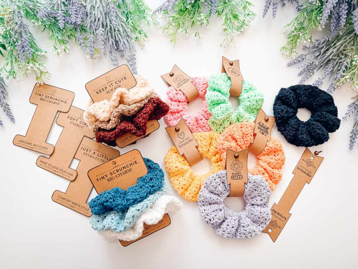 Crocheted scrunchies in various colors are arranged with brown cardboard tags on a white surface, surrounded by sprigs of purple and green artificial flowers—perfect accessories to pair with your favorite crochet nesting trays pattern.