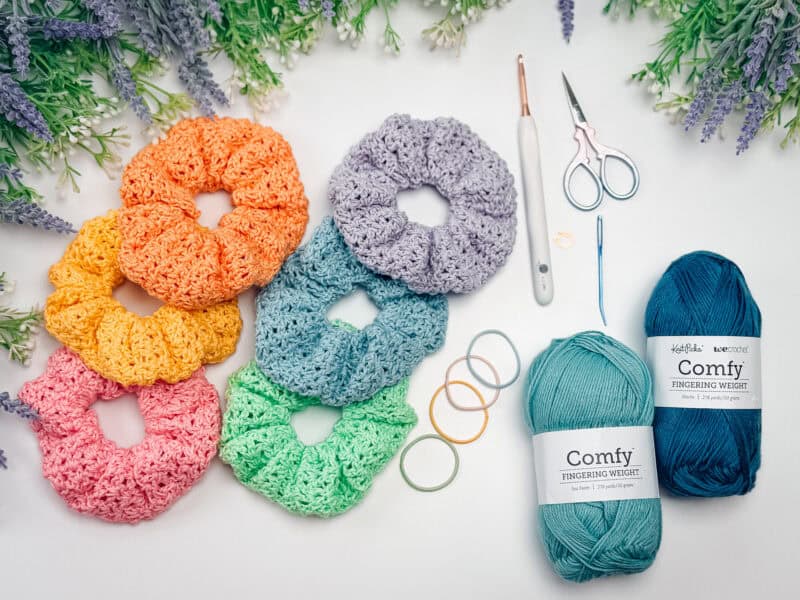 Six crocheted scrunchies in various colors, two skeins of teal yarn, scissors, a crochet hook, and stitch markers are arranged on a white surface with floral accents—perfect inspiration for your next crochet nesting trays pattern.