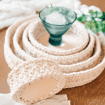 A set of round crochet storage baskets, made using a crochet nesting trays pattern, sit nested on a wooden surface with decorative glass and fabric nearby. Text reads: “Stylish Crochet Storage Ideas For Your Home.”.