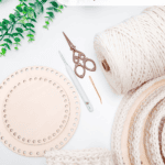 Crochet supplies including yarn, wooden circles with holes, scissors, a crochet hook, and a finished crochet nesting trays pattern are laid out on a white surface with green leaves overhead.