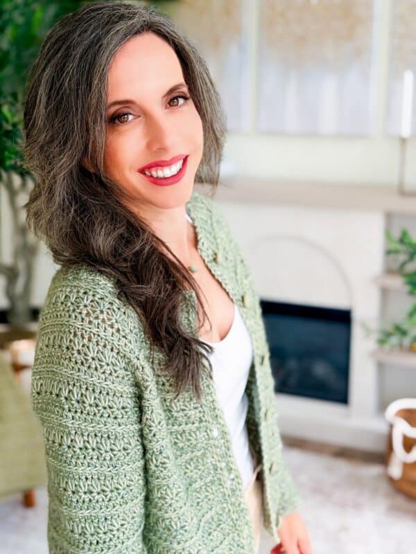 A woman with long, wavy graying hair smiles at the camera. She wears a light green Petal Loop Stitch Crochet Cardigan over a white top, standing in a bright, modern living room.
