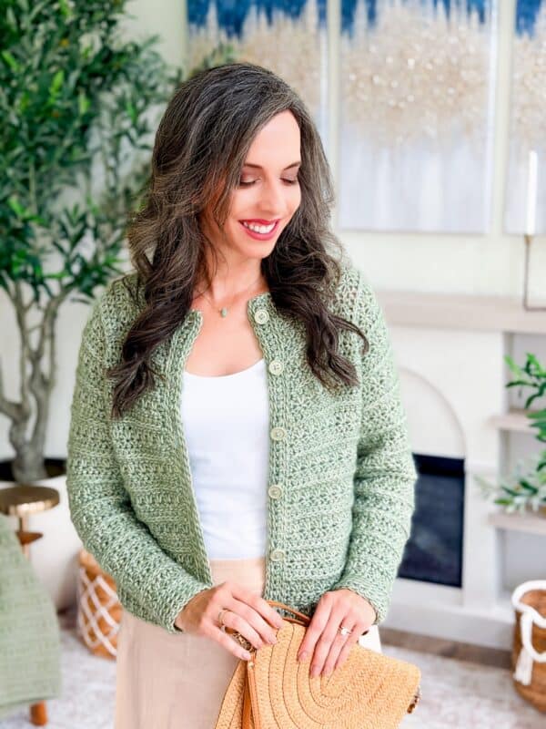 Woman with long, wavy brown hair wearing a light green crochet cardigan pattern over a white top, holding a woven bag, and standing indoors near plants and artwork.