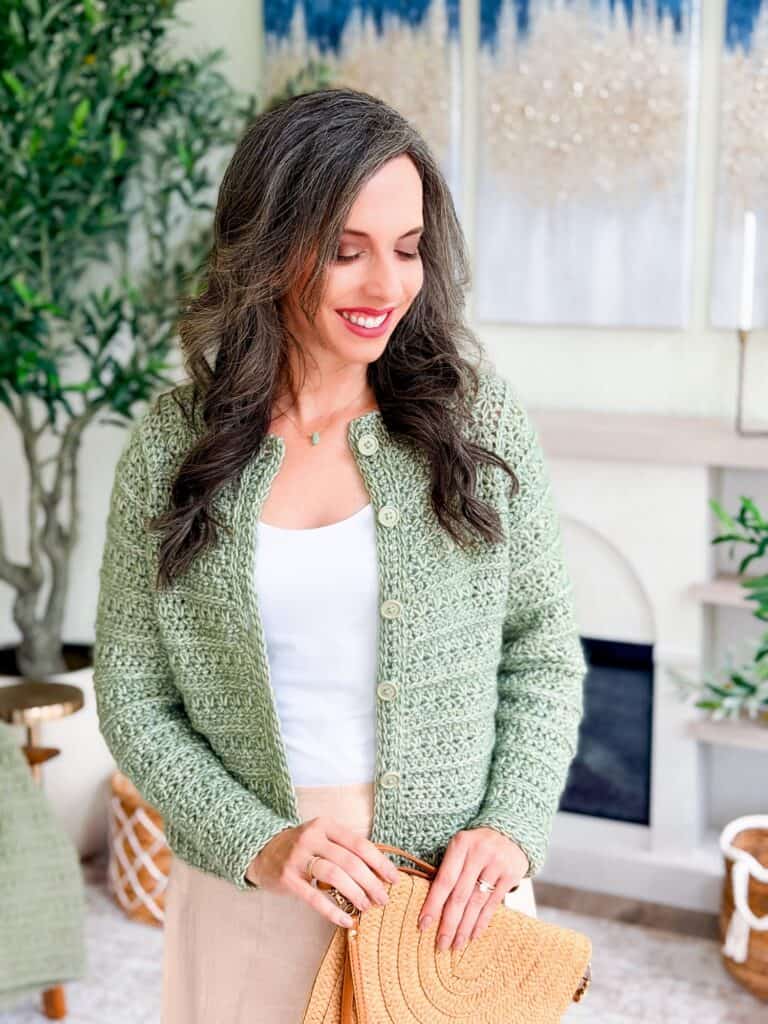 Woman with long, wavy brown hair wearing a light green crochet cardigan pattern over a white top, holding a woven bag, and standing indoors near plants and artwork.