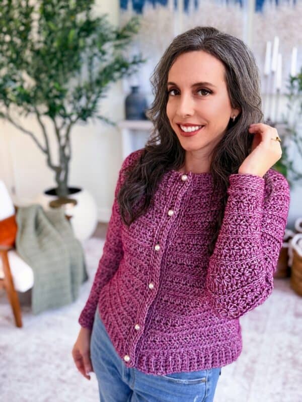 A woman with long dark hair wears a cardigan from the petal loop stitch crochet cardigan pattern in purple with pearl buttons and blue jeans, standing indoors near plants and home decor.
