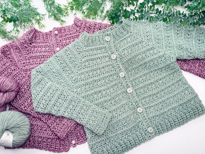 Two hand-crocheted Petal Loop Stitch Crochet Cardigans, one light green and one mauve, are laid flat with matching yarn balls and green leafy plants in the background.