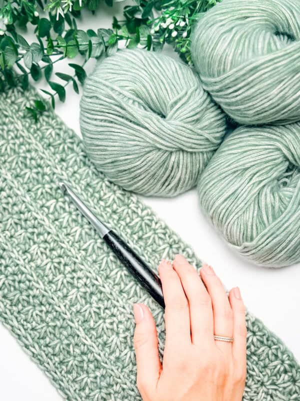 A hand holding a crochet hook rests on a partially crocheted green crochet cardigan, with green yarn balls and artificial leaves nearby.