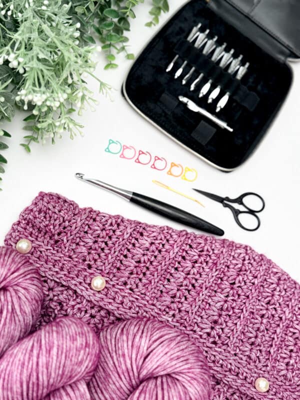 A crochet project from the Petal Loop Stitch Crochet Cardigan Pattern with pink yarn, a crochet hook, scissors, needle, stitch markers, and a case of hooks, set on a white surface beside green foliage.