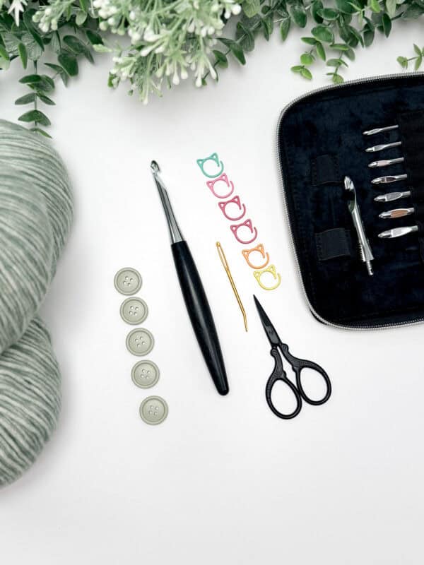 Crochet supplies for the crochet cardigan pattern including yarn, buttons, stitch markers, scissors, a crochet hook, a yarn needle, and a case of crochet hooks are arranged on a white surface with greenery—perfect for crafting your next Wheat Stitch Crochet Sweater.