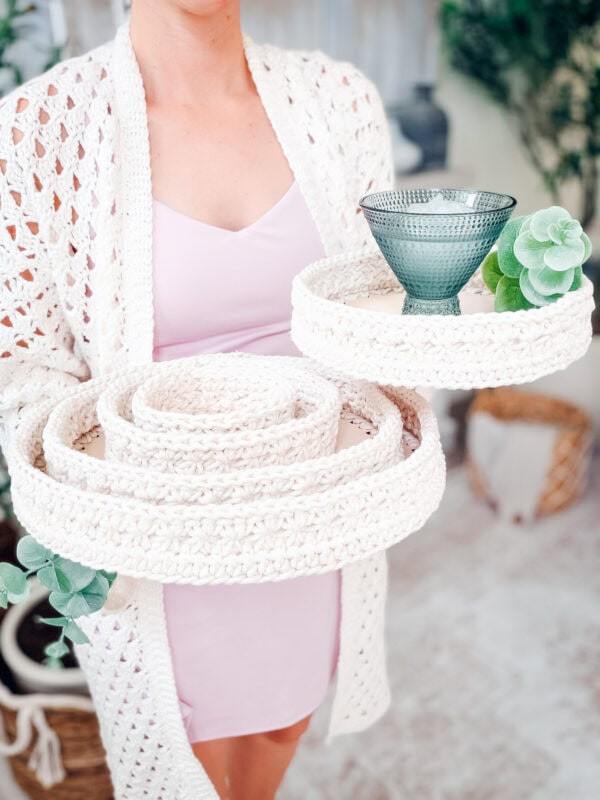 A person in a light pink dress and white crochet cardigan holds a set of crochet nesting trays, made using a stylish crochet nesting trays pattern, one tray holding a green glass and a small faux succulent.