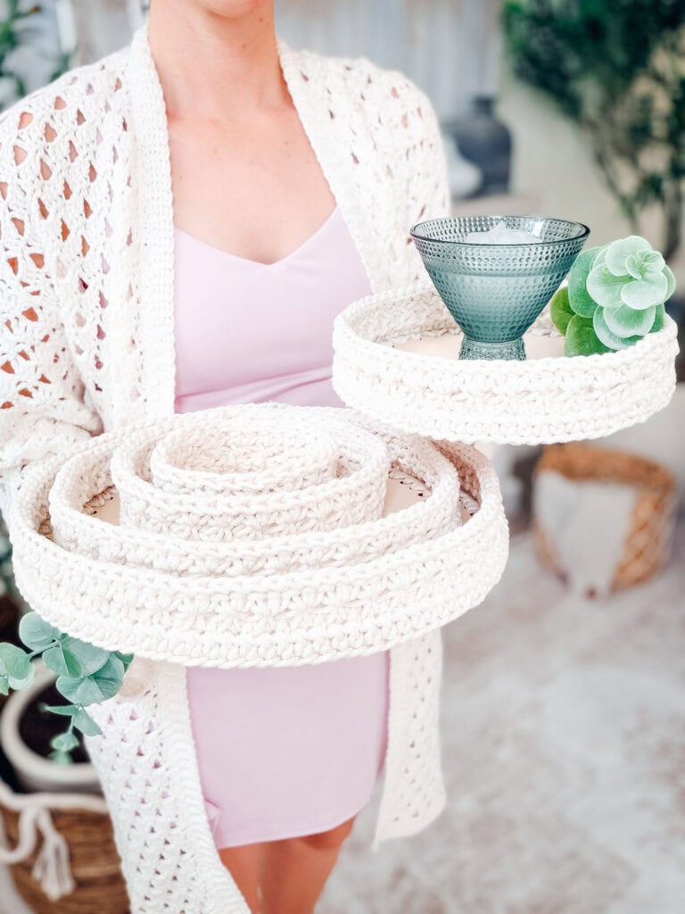 A person in a light pink dress and white crochet cardigan holds a set of crochet nesting trays, made using a stylish crochet nesting trays pattern, one tray holding a green glass and a small faux succulent.