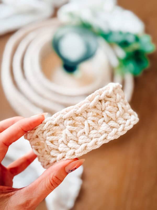 A hand holds a small, rectangular piece from a crochet nesting trays pattern, with a coiled rope and a green plant softly blurred in the background.