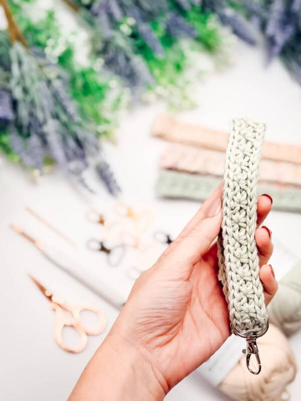 A hand holding a crocheted keychain wristlet; scissors, crochet hooks, yarn, and a crochet nesting trays pattern are visible among other crocheted items in the blurred background.