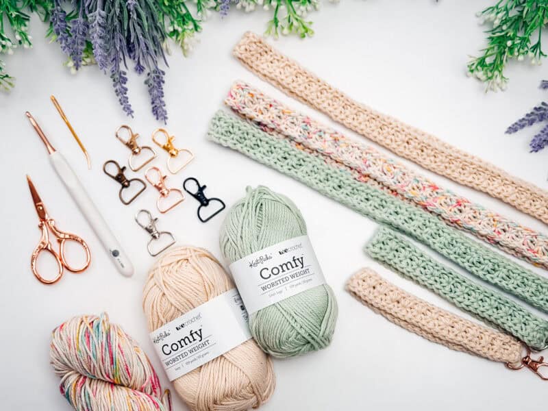 Three skeins of yarn, crochet hooks, scissors, metal clips, and finished crochet straps&mdash;perfect supplies for your next project&mdash;are arranged on a white surface with greenery in the corners, inspiring ideas for a crochet nesting trays pattern.