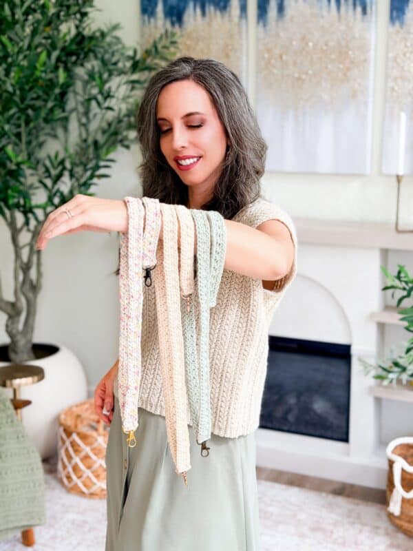 Woman with long brown hair stands indoors, smiling as she holds several crocheted keychain wristlets draped over her raised arm. She wears a cream knit top and green skirt, perfectly matching her crochet nesting trays pattern creations.