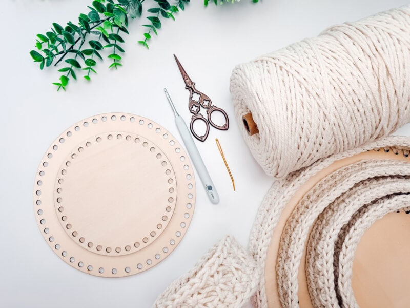 Round wooden base with holes, crochet hook, scissors, yarn needle, large ball of cord, and crocheted Petal Loop Stitch Blanket pieces on a white surface with a green plant in the corner.