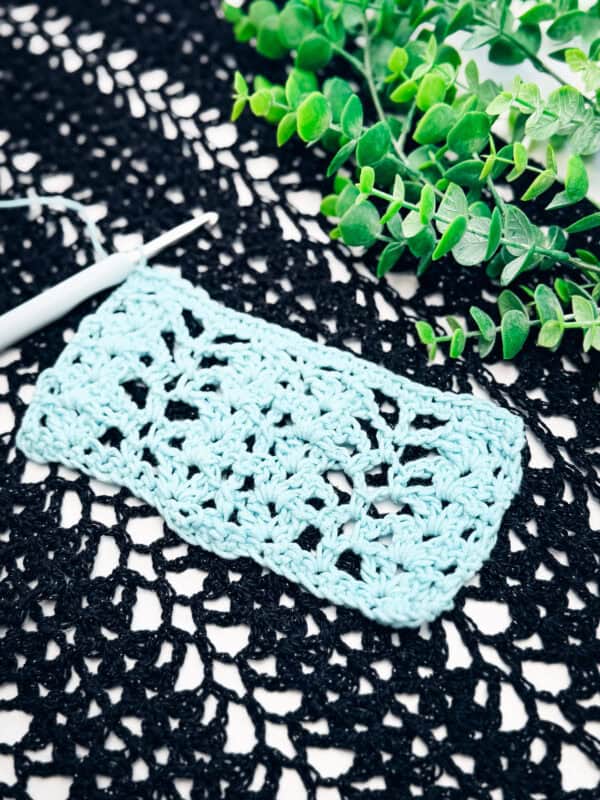 A blue crochet swatch with a lacy pattern rests on a black crocheted fabric, next to a white crochet hook and green artificial plants.