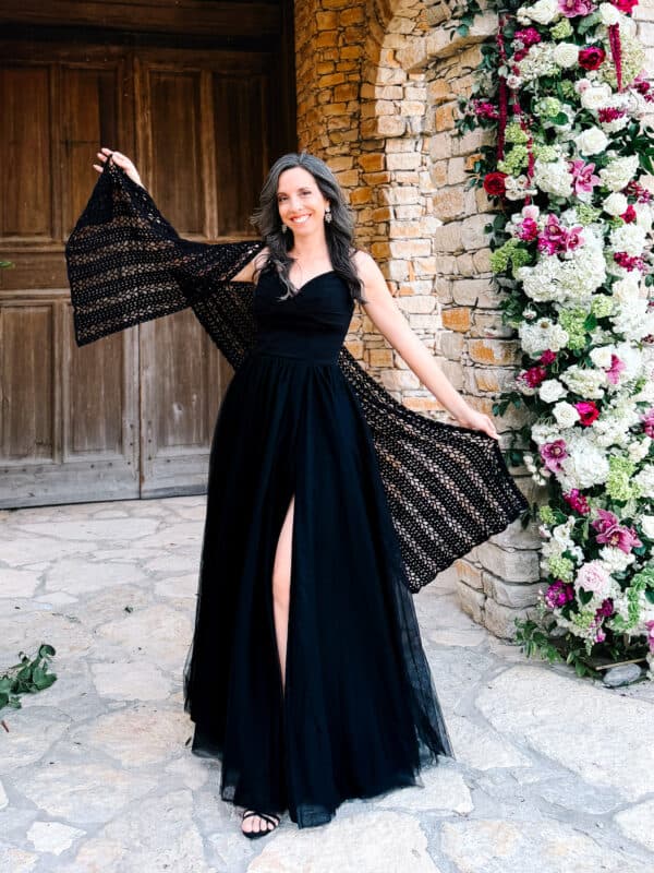 Woman in a long black dress poses outdoors on stone pavement, holding out a lacy shawl reminiscent of the Willow Mesh Stitch crochet tutorial, with a floral arrangement and wooden doors in the background.