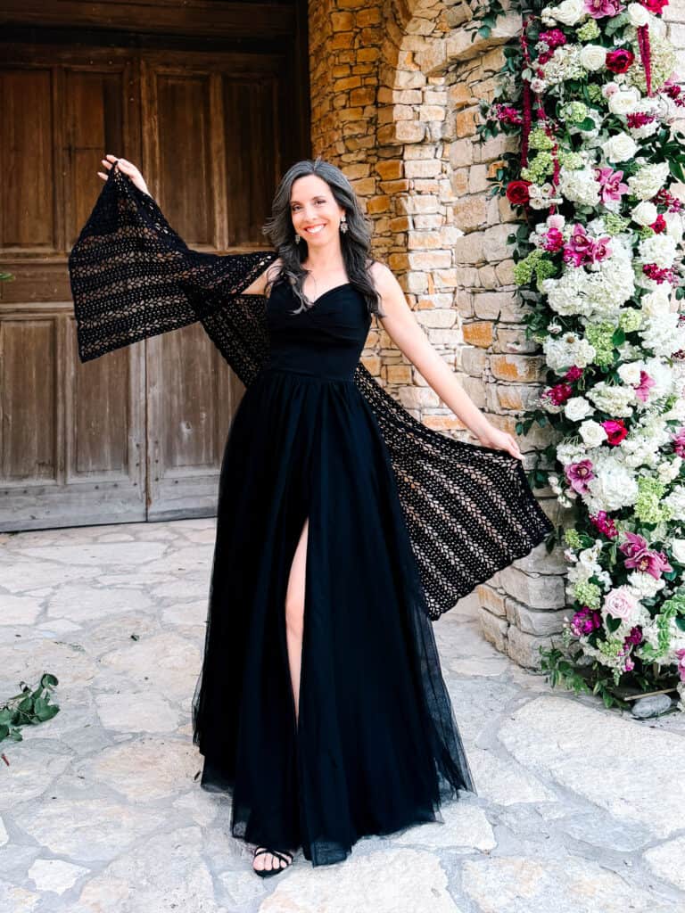 Woman in a long black dress poses outdoors on stone pavement, holding out a lacy shawl reminiscent of the Willow Mesh Stitch crochet tutorial, with a floral arrangement and wooden doors in the background.