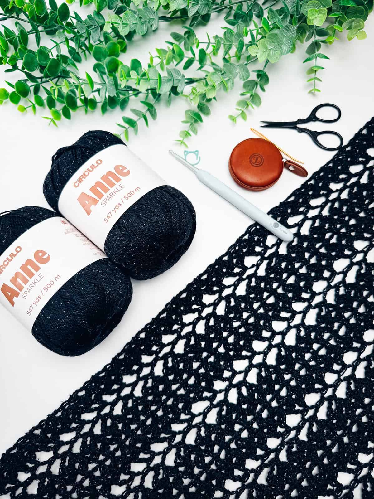 Two black yarn skeins, a crochet hook, scissors, measuring tape, a black lace crochet piece, and green faux leaves arranged on a white surface.