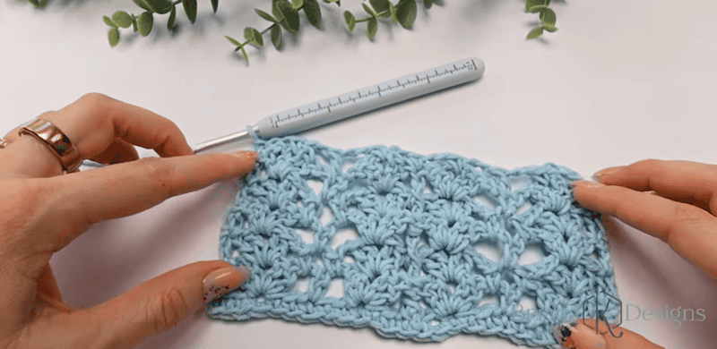 A pair of hands holds a light blue crocheted swatch with a lacy pattern; a crochet hook and green leaves are in the background.