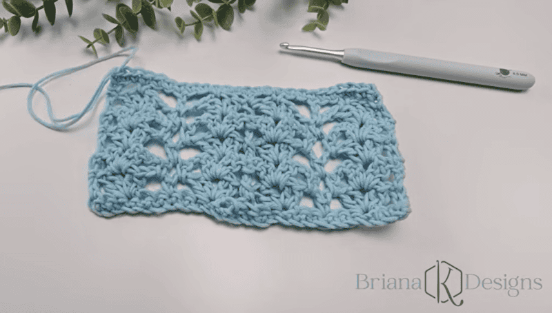 A light blue crochet swatch with a textured, lacy pattern lies next to a crochet hook on a white surface, with green leaves in the background.