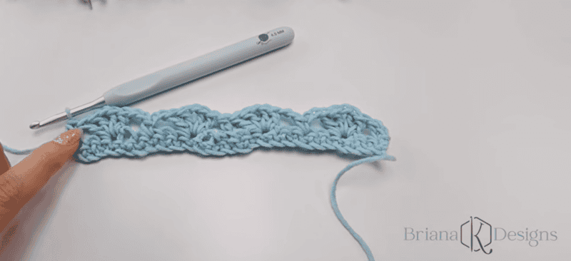 A hand points to a blue crochet scallop pattern in progress next to a grey crochet hook on a white surface. "Briana K Designs" is visible in the bottom right corner.