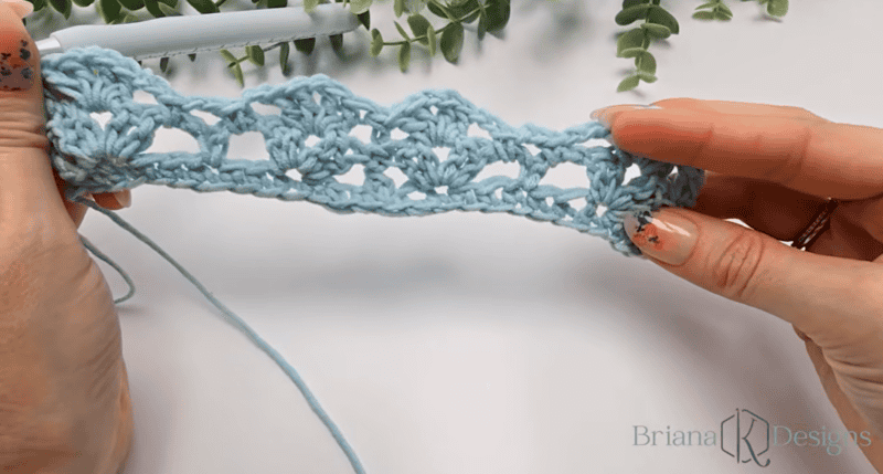 Hands holding a light blue crochet project with a shell-like openwork pattern, a crochet hook, and greenery in the background. Briana K Designs logo is visible in the lower right corner.