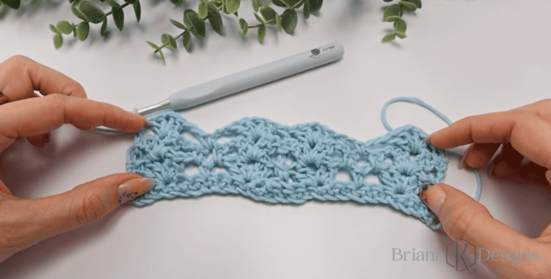 Two hands hold a crocheted light blue swatch with an open, lacy pattern. A crochet hook and green plant leaves are in the background.