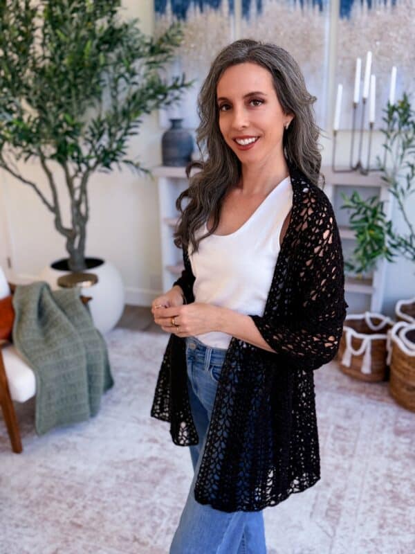 A woman with long gray hair, wearing a white top, jeans, and a black crocheted cardigan, stands indoors with plants and woven baskets in the background—her style perfect for showcasing the Willow Mesh Stitch crochet tutorial.