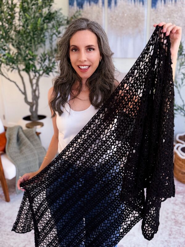 A woman with long gray hair wearing a white tank top holds up a large, black, lacy crocheted shawl indoors.