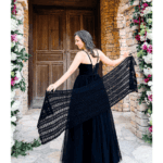 Woman in a black dress stands in front of a rustic wooden door, holding out a long, Willow Mesh Stitch crochet wrap; floral arrangements frame the doorway.