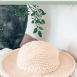 A beige crochet sun hat with a braided band displayed on a wooden surface near a plant, advertising sizes from newborn to adult large.