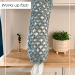 A crocheted bag organizer filled with plastic bags hangs in a kitchen; a dog sniffs a bag at the bottom, and a potted plant sits on the counter nearby.