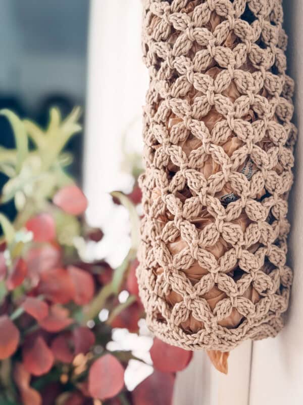 Close-up of a beige crocheted bag holder containing plastic bags, hanging on a wall near a plant with green and reddish leaves.