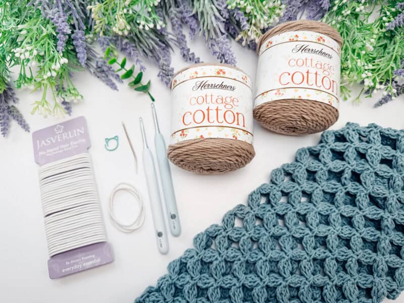 Yarn, crochet hooks, tapestry needles, elastic cord, and a crocheted fabric square are arranged on a table with artificial flowers in the background.