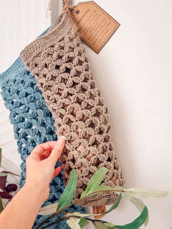 A hand holds a beige crocheted bag hanging on a wall beside a blue crocheted bag, with a brown tag attached and green leaves in the foreground.
