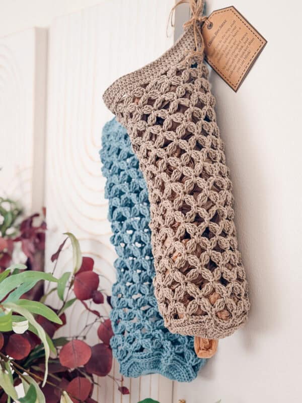Two crocheted mesh bags, one beige and one blue, hang on a wall. A brown tag is attached to the beige bag. Greenery and purple leaves are visible in the lower left corner.
