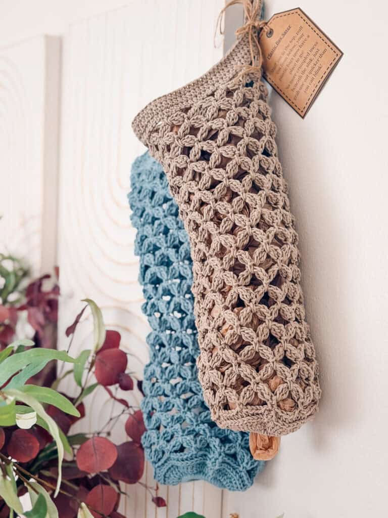 Two crocheted mesh bags, one beige and one blue, hang on a wall. A brown tag is attached to the beige bag. Greenery and purple leaves are visible in the lower left corner.